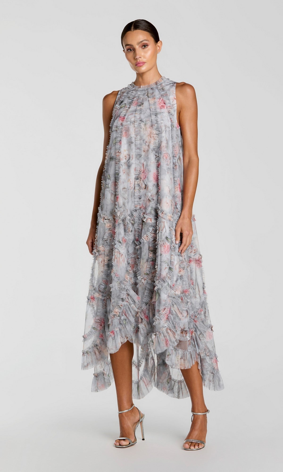 A woman stands against a plain background wearing the Midi Party Dress: Mac Duggal 8430 in Pearl Grey—a sleeveless, high-neck, flowy floral cocktail dress featuring pink patterns and a ruffled hem—paired with silver high-heeled sandals.